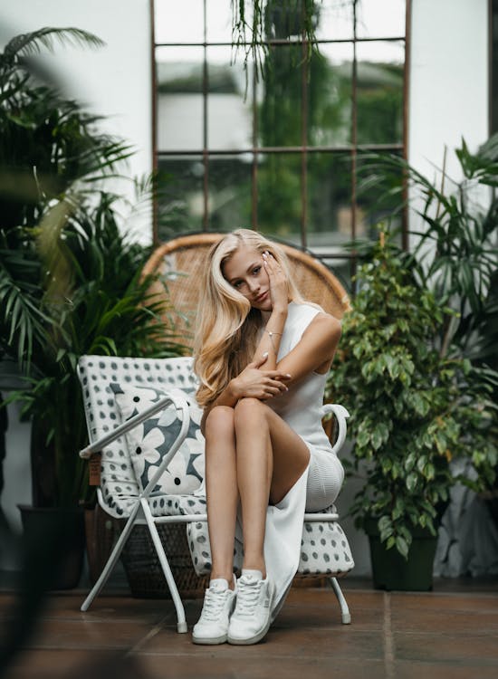 Young Woman in a White Dress and White Sneakers Sitting on a Chair between Plants