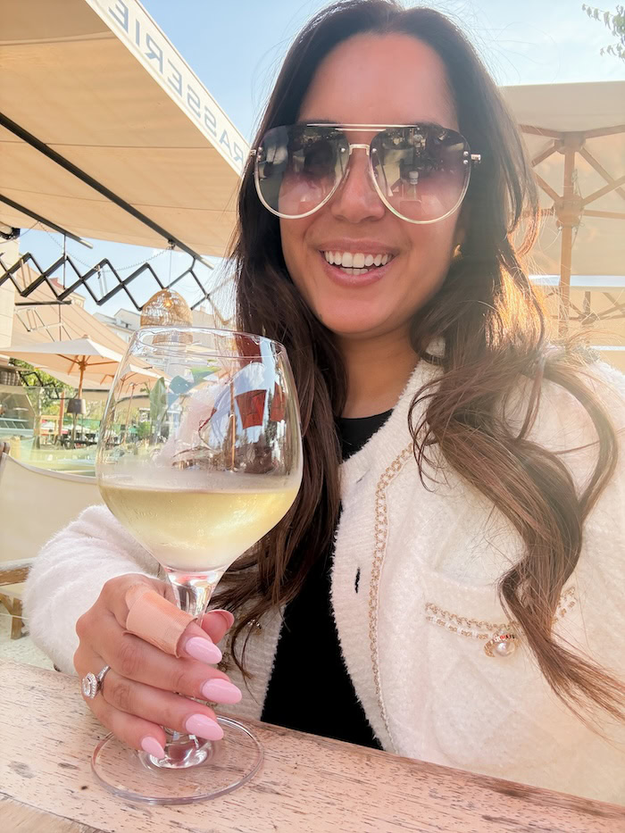Woman smiling in sunglasses holding a glass of white wine at an outdoor cafe in Cannes, wearing a white tweed cardigan