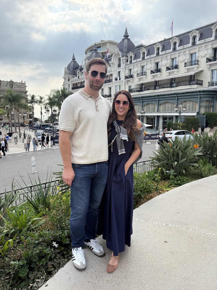 Couple posing in Monaco in front of Hotel de Paris, woman wearing navy tank dress with striped sweater tied over shoulders